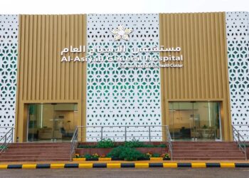 Exterior of Al-Asyah General Hospital with a modern facade. The building features a white perforated pattern with golden accents and Arabic text above the entrance. Two glass doors are visible, flanked by small sets of stairs and a striped path.