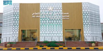Exterior of Al-Asyah General Hospital with a modern facade. The building features a white perforated pattern with golden accents and Arabic text above the entrance. Two glass doors are visible, flanked by small sets of stairs and a striped path.