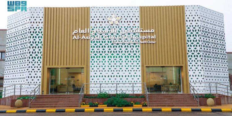 Exterior of Al-Asyah General Hospital with a modern facade. The building features a white perforated pattern with golden accents and Arabic text above the entrance. Two glass doors are visible, flanked by small sets of stairs and a striped path.